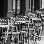 A line of wicker patio chairs and glass bistro tables on a streetside patio.